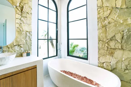 Luxurious modern bathroom with textured stone walls, arched black-framed windows overlooking tropical palms, freestanding white bathtub filled with red petals, and sleek vanity sink. - Bali Villas